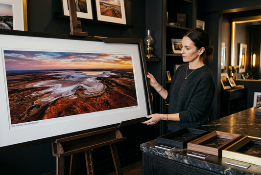 Woman examining a large framed aerial print of Kati Thanda-Lake Eyre at sunset in a gallery showroom with frame samples.
