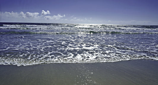 A scenic photograph of ocean waves with white foam rolling onto a sandy beach under a blue sky with light clouds.