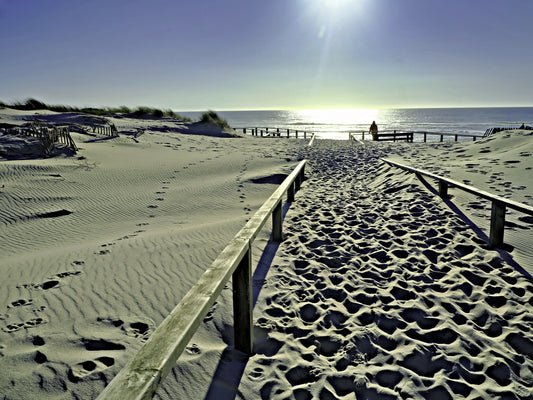 A sandy beach path with wooden railings leading toward the ocean under a bright sun, with footprints in the sand and a small person standing near the shoreline.
